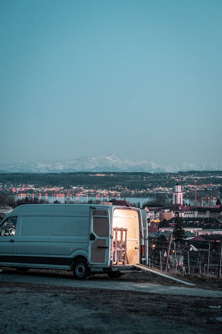 A silver van parked on a slightly elevated area during dusk or early evening, with its rear doors open revealing furniture and supplies positioned inside for loading or unloading as part of a home rel