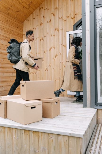 The image depicts a wooden outdoor patio area attached to a house, with vertical wooden paneling on the walls and a light-colored wooden decking floor. On the patio, there are three cardboard boxes st