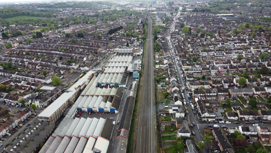 An aerial view of a residential area showing densely packed houses, streets, and greenery, with a large rail depot or warehouse facility featuring long, white, peaked roofs situated adjacent to a railway line running through the centre of the image. The railway tracks extend into the distance, bordered by urban and suburban landscape. The scene captures the urban environment surrounding Colliers Wood, with some vehicles visible on the roads. This image supports content related to home relocation and furniture transport services offered by Man with Van Colliers Wood, illustrating the logistical context of domestic removal and moving processes near Colliers Wood Station, with the railway and warehouse elements emphasizing transportation infrastructure involved in efficient house removals.