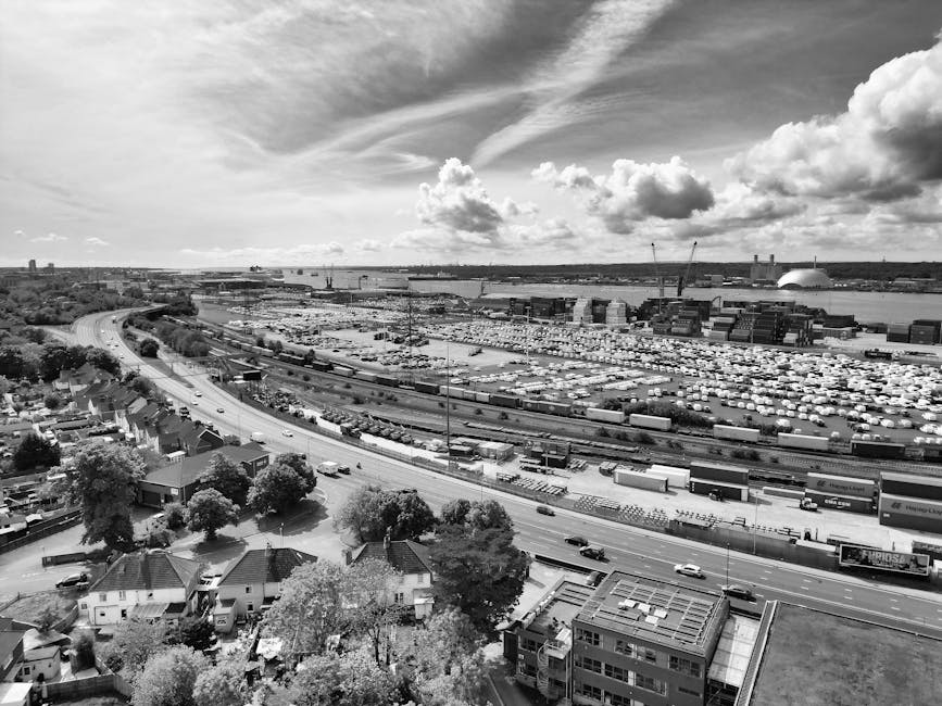 A wide aerial view in black and white showing a large industrial port area with numerous shipping containers, cranes, and a docked cargo ship along a waterway. In the foreground, a residential neighborhood with houses, trees, and streets is visible, leading towards a busy road running parallel to the port. The scene includes a partly cloudy sky with high-altitude clouds and scattered cumulus clouds, and distant urban infrastructure across the horizon. The image captures a mix of urban residential and industrial zones, relevant to house removals and transportation logistics, as provided by Man with Van Colliers Wood.